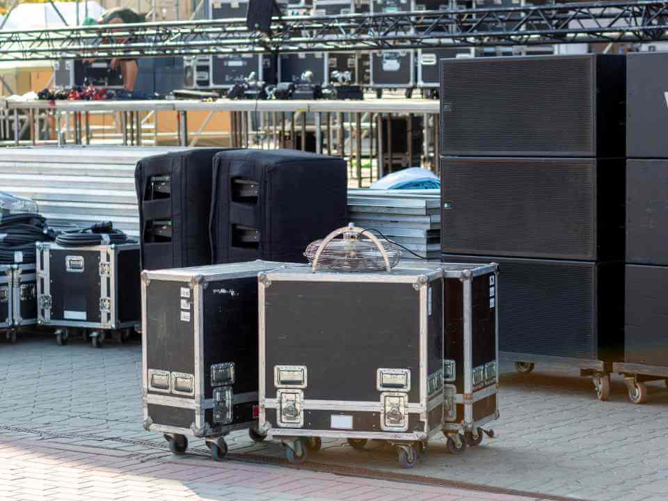 Stacked flight cases, loudspeakers, and coiled cables arranged on a paved outdoor surface near a stage setup.